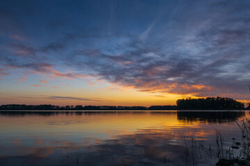 A peaceful sunset over a tranquil lake near Alkmaar, Netherlands, as the sky fades to soft hues of orange and pink, reflecting gently on the calm waters and surrounding countryside