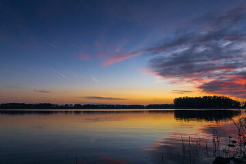 A peaceful sunset over a tranquil lake near Alkmaar, Netherlands, as the sky fades to soft hues of orange and pink, reflecting gently on the calm waters and surrounding countryside