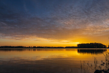 A peaceful sunset over a tranquil lake near Alkmaar, Netherlands, as the sky fades to soft hues of orange and pink, reflecting gently on the calm waters and surrounding countryside
