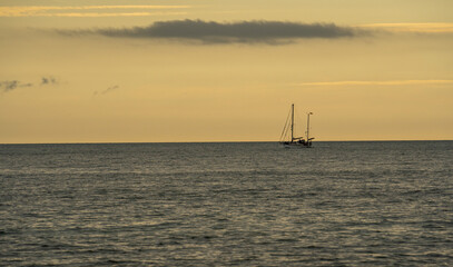 The silhouette of a two-masted sailboat on the calm Baltic Sea against the warm, golden glow of a...