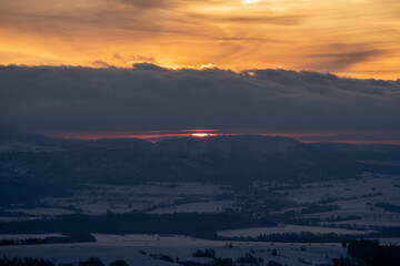 The last sliver of the setting sun disappears behind a dark mountain ridge in a dramatic, snowy winter landscape. Picture taken on a holiday trip to Gniewoszów, Poland with my Sony a7III