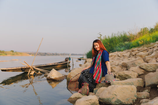 With a warm smile, a young woman in traditional attire sits on the rocky bank of the Mekong River. A friendly and inviting portrait of a person deeply connected to her culture and home.