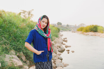 A confident young woman in traditional Isaan attire, with a scarf covering her head, stands by the...