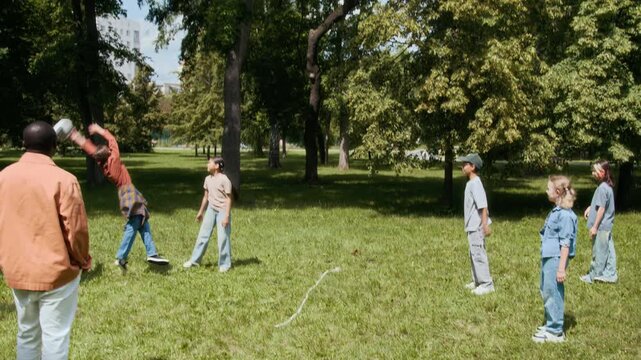 Black young male teacher watching six multiethnic gen alpha children playing throwball on summer day in park