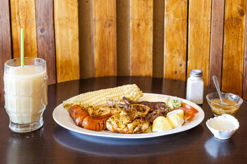 Ecuadorian roast with chicken, beef, and sausage, served with corn on the cob, fresh cheese, potatoes with sauce, and a tomato-lettuce salad. Accompanied by an oatmeal colada with naranjilla.
