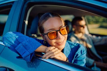 Carefree woman wearing sunglasses leaning out of car window enjoying road trip with her friend driving