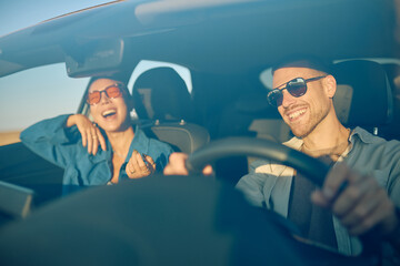 Happy couple wearing sunglasses, driving joyfully and laughing together during a picturesque road trip at sunset, embracing freedom and adventure