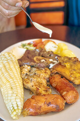 Pouring homemade mayonnaise over an Ecuadorian barbecue, with a mix of meats like chicken, chorizo, and beef, accompanied by corn, fresh cheese, potatoes in salsa, and a tomato and lettuce salad.