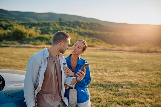 Young multi-ethnic couple using smartphone next to their car during a road trip at sunset, enjoying summer holidays together