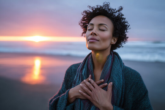 Peaceful woman enjoying a serene sunrise on the beach with eyes closed and hands over heart, embracing calm and mindfulness