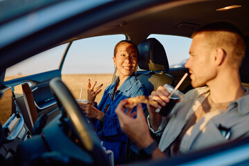 Happy couple enjoying pizza and sipping soda inside their car, taking a delightful break during a summer road trip at sunset