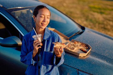 Asian woman enjoying pizza and sipping soda while taking a break near her car during a picturesque sunset on a road trip