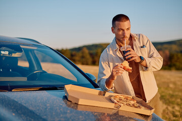 Young man enjoying pizza and soda during a break on a road trip, using car hood as table in a countryside setting