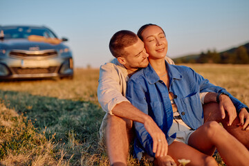 Young multi-ethnic couple enjoying a romantic moment sitting in a field at sunset during a road trip with their car parked in the background