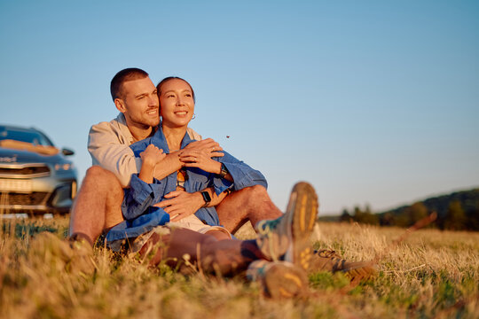 Young couple enjoying a romantic moment during a road trip, embracing in a field at sunset near their parked car