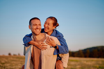 Interracial couple having fun in a meadow at sunset, woman piggybacking on man, laughing and enjoying time together