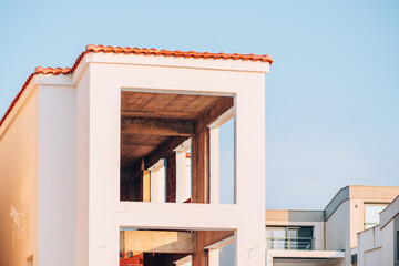 Minimalist view of an unfinished modern building with open concrete interior, white exterior walls, and terracotta roof tiles under clear blue sky.