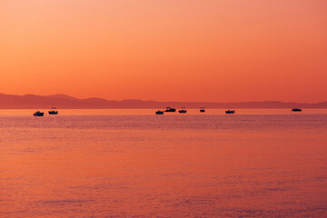 Fototapeta premium Silhouetted boats floating on calm sea during vibrant orange sunrise with distant mountains and smooth water reflections.