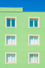Minimalist facade of a green building with six identical white-framed windows under clear blue sky, captured in bright sunlight.