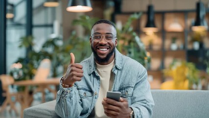 A happy bearded man with glasses holds a smartphone and gives a thumbs-up smiling warmly in a modern indoor setting - Powered by Adobe