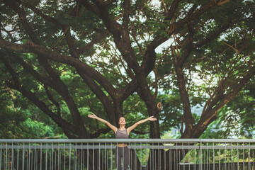 Happy Asian woman standing on a bridge in sportswear with arms open wide after a successful run at the outdoor park, surrounded by green trees, smiling and relaxing with a sense of freedom