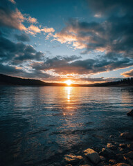 The sun sets over a peaceful lake, casting colorful reflections on the water's surface as clouds drift above