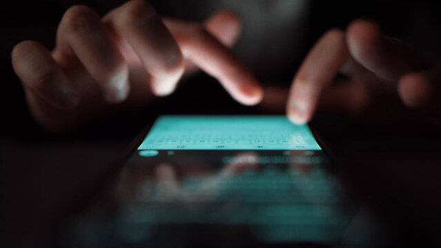 A close-up view captures hands interacting with a smartphone screen in a dimly lit setting, highlighting modern communication and technology usage in daily routines. Xenic