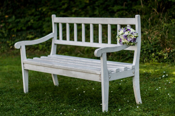 Fototapeta premium White wooden bench holding wedding bouquet in garden setting