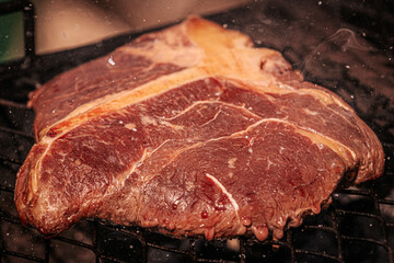 Close-up of a thick raw T-bone steak sizzling on a grill with visible marbling and seasoning droplets in the air, capturing the moment of flavoring.