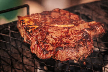Two juicy bone-in steaks sizzling on a hot grill, one being flipped with metal tongs. Rich grill marks and charred edges highlight the heat and texture.