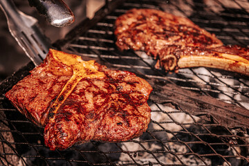 Two juicy bone-in steaks sizzling on a hot grill, one being flipped with metal tongs. Rich grill marks and charred edges highlight the heat and texture.