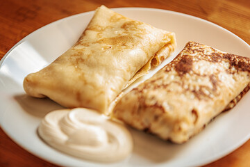Two golden-brown stuffed crepes served on a white plate with a swirl of sour cream. The texture is soft and slightly crispy with a warm, homemade appeal.