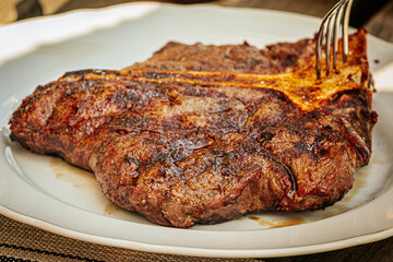 Close-up of a juicy grilled T-bone steak on a white plate, being sliced with a fork and knife. Cooked to perfection with visible grill marks and seared crust.