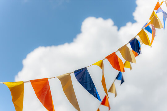 Vibrant orange, yellow, blue, and beige pennant flags flutter in the wind under a sunny sky with fluffy white clouds, creating a festive and cheerful mood. Includes copy space.