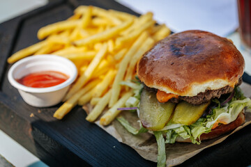 Juicy cheeseburger with pickles, lettuce, and sauce served on a toasted bun, paired with crispy French fries and a side of ketchup on a black wooden tray.