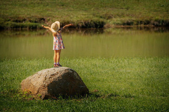 A young girl in a floral dress and sunhat stands on a large rock by a pond, arms outstretched in joy. The serene natural setting suggests freedom and childhood wonder.