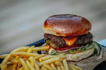 Juicy cheeseburger with pickles, lettuce, and sauce served on a toasted bun, paired with crispy French fries and a side of ketchup on a black wooden tray.