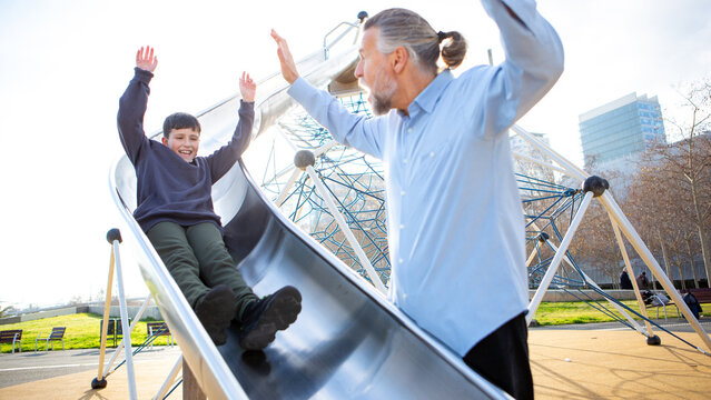 Boy sliding down playground slide for high-five with grandpa