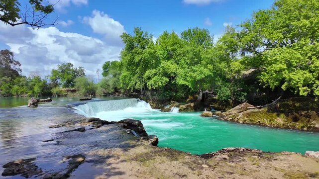 Manavgat waterfall captured in serene natural environment with wide stream of turquoise water flowing over rocks beneath sunny blue skies in picturesque national park, slow motion