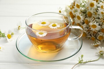 Aromatic tea in glass cup and chamomile flowers on white wooden table, closeup