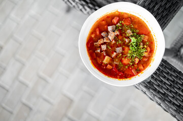 Homemade beef and vegetable soup served in a white bowl on a wicker table, featuring fresh herbs and colorful ingredients like potatoes, carrots, and tomatoes.