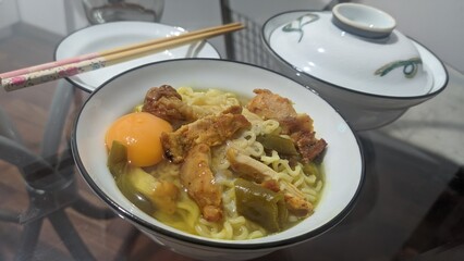 Homemade instant ramen noodle soup in a white bowl with chicken, raw egg yolk, and pickled vegetables. Traditional Asian meal with chopsticks on a glass table. Comfort food concept for dinner or lunch