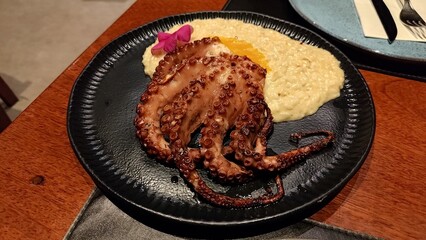 Gourmet grilled octopus tentacle served with creamy risotto, garnished with an edible flower and orange zest on a stylish black plate. A fine dining seafood dish on a wooden restaurant table.
