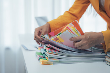 A businesswoman is working with a large pile of paperwork, searching for unfinished documents, information on piles of documents on her desk, and checking financial documents amidst her busy workload.