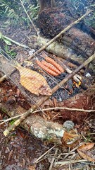 Rustic outdoor barbecue with a whole fish and sausages grilling on a rack over a campfire. Primitive cooking in a forest setting, showing a survival and adventure lifestyle concept.