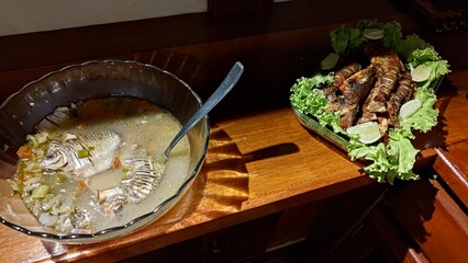 A complete seafood meal featuring two traditional preparations. A hearty fish soup in a glass bowl alongside crispy fried fish on a platter, both garnished and served on a classic wooden table.