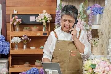 Asian senior female florist multitasking during workday in flower shop, making phone call while...