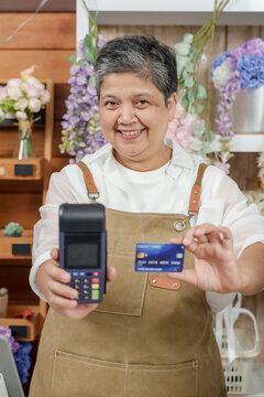 Asian middleaged female florist wearing apron holding credit card and card reader in flower shop smiling for contactless payment service concept with fresh blossoms and professional retail setup