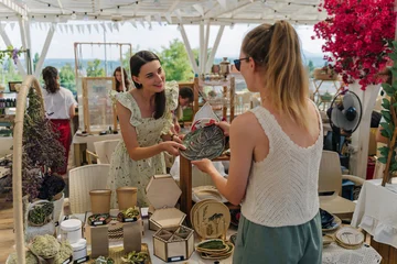 Seller Showing Handmade Ceramic Plate to Buyer at Summer Outdoor Artisan Market. Handmade Ceramics with Leaf, Floral, and Fruit Ornamentation © igishevamaria