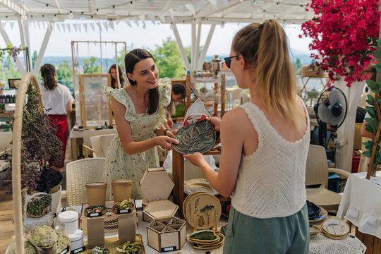 Seller Showing Handmade Ceramic Plate to Buyer at Summer Outdoor Artisan Market. Handmade Ceramics with Leaf, Floral, and Fruit Ornamentation